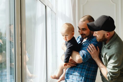 Two men holding a baby, looking out a bright window, smiling and enjoying the view, as they cherish their latest key initiative in the journey of parenthood.